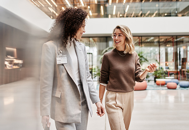 Zwei Frauen, eine im grauen Blazer mit Namensschild und die andere im braunen Pullover, gehen lächelnd in einem modern eingerichteten Bürogebäude mit vielen Fenstern.