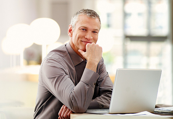 Ein Mann sitzt an einem Schreibtisch, stützt seinen Kopf auf seine Hand und lächelt, während er vor einem geöffneten Laptop in einem modernen, hellen Büro sitzt und über die Interflex Managed Services Cloud Services nachdenkt.