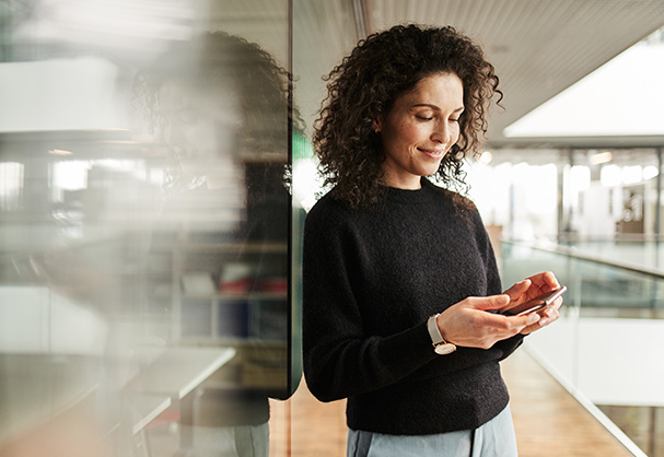 Eine Frau mit lockigem Haar hält ein Smartphone und lächelt. Sie steht in einem modernen, hellen Büro mit Glaswänden und Holzboden, bei Tageslicht durchflutet.