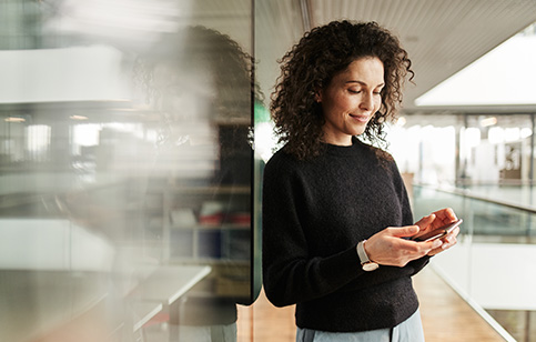 Eine Frau mit lockigem Haar hält ein Smartphone und lächelt. Sie steht in einem modernen, hellen Büro mit Glaswänden und Holzboden, bei Tageslicht durchflutet.