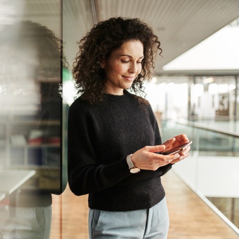 Vrouw leunt tegen het glazen raam, glimlachend en kijkend naar haar smartphone waarop ze haar tijdregistratie invult