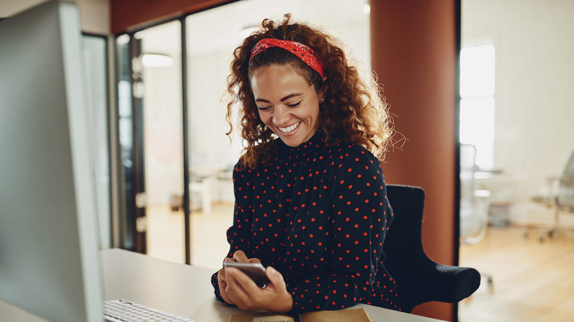 Woman sitting at her desk, smiling and looking at her smartphone